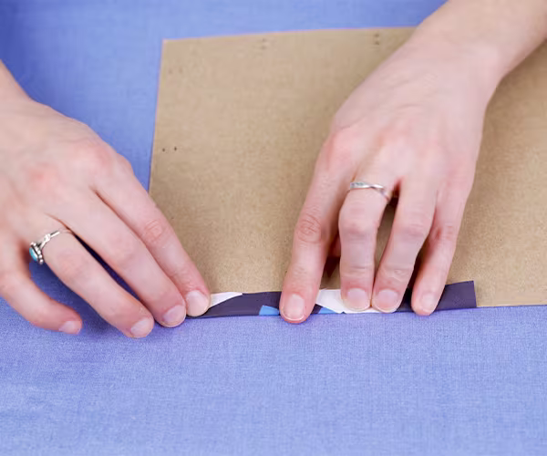 Folding a small section of a fabric over a piece of cardboard to create a seam. The cardboard is acting like a seam template.
