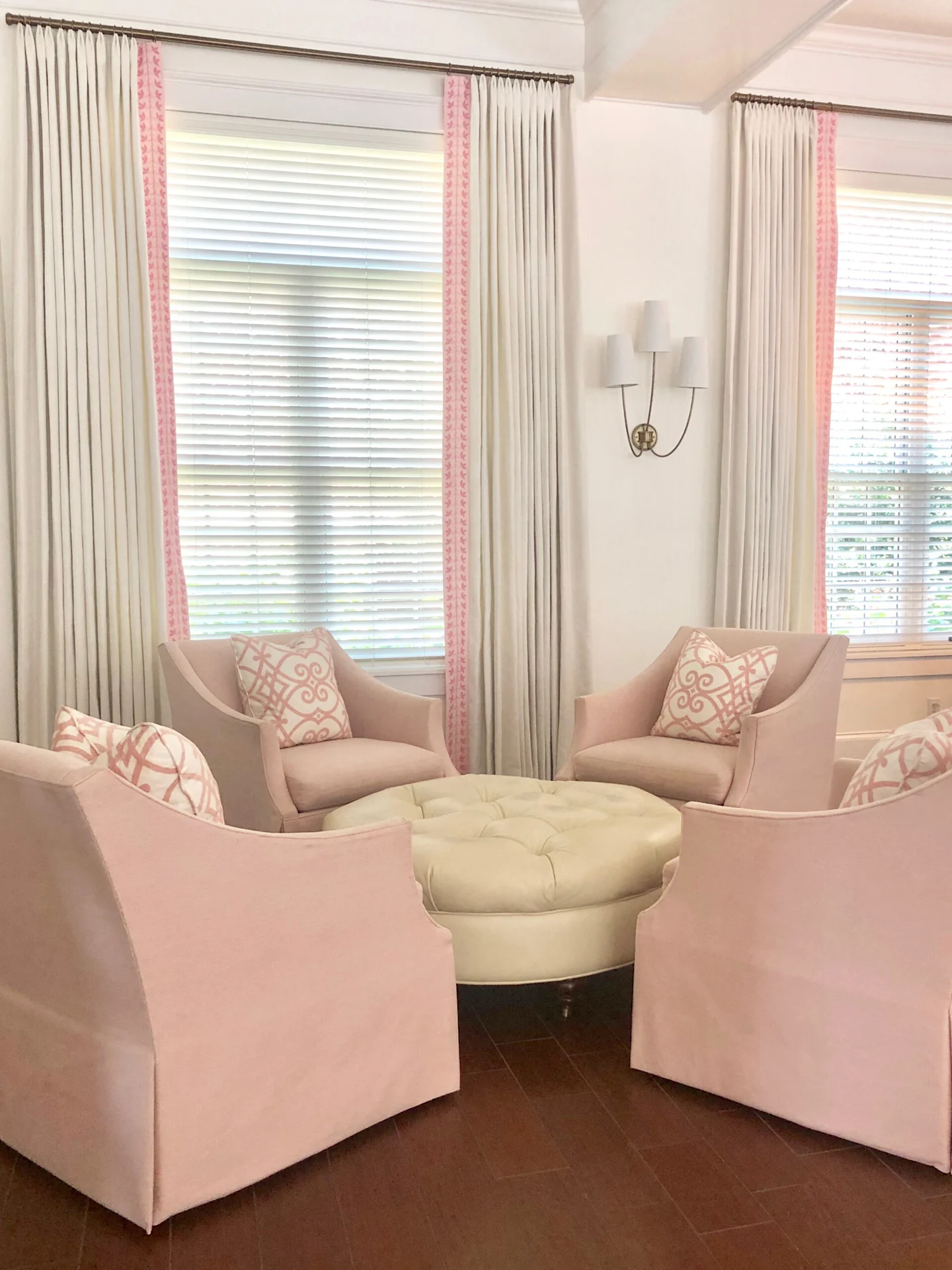 Four light pink chairs sit around a round white table. The pillows on the chairs are cream with a light pink lacy design. The white curtains behind two of the chairs have banding on the fabric with a light pink background with darker pink leaves.