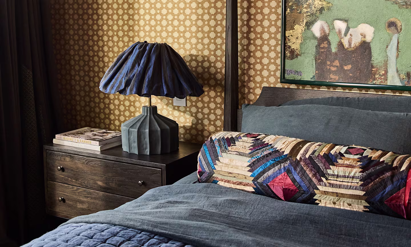 A cozy, design-forward bedroom featuring geometric wallpaper and deep blue bedding. A sculptural table lamp with a gathered navy shade sits beside a stack of books, while a colorful quilted accent pillow adds texture and personality to the space..
