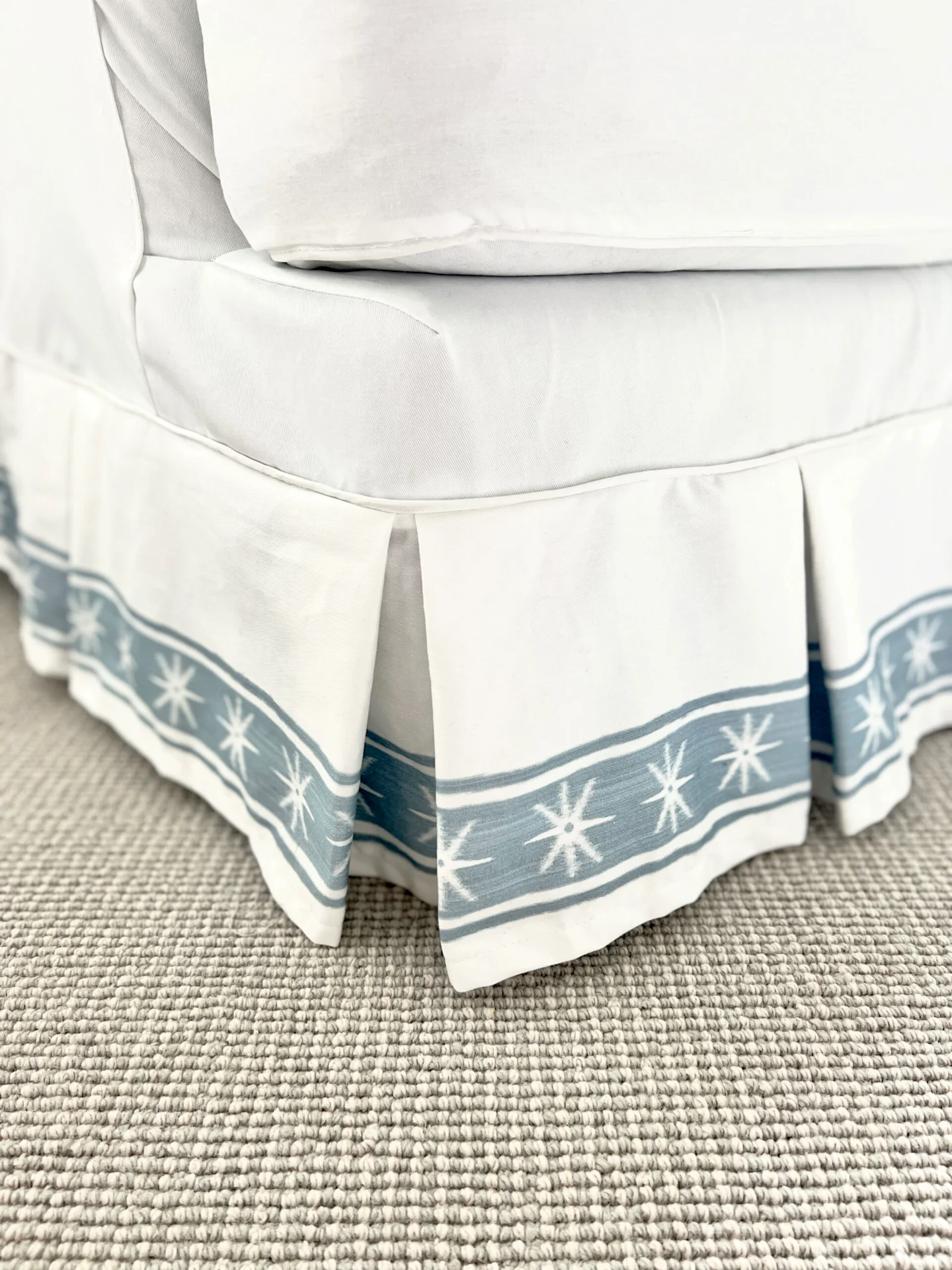 A white chair has a strip of white fabric with a vertical thick blue stripe with white stars and a thin blue stripe on either side of each thicker stripe along the bottom edge of its dust ruffle fabric.