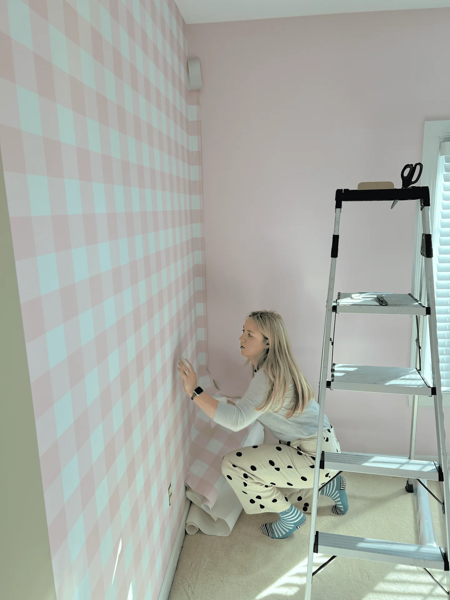 A woman is bent down as she installs wallpaper with a pink-and-white gingham design on a wall making sure the pattern lines up.