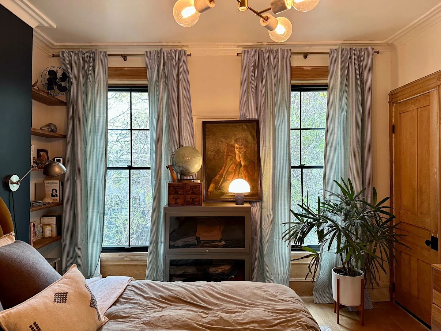 Blue-and-white striped curtains frame two large windows at the back of a bedroom. A bed with a tan comforter is in the foreground. A small bookcase sits between the windows; it has a small dome lamp, a painting of a man looking pensive and a globe on top 