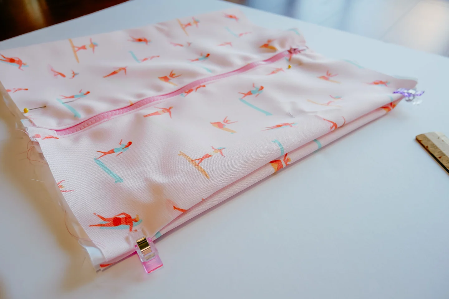 A fabric rectangle featuring a design with small female surfers surfing through a pink background lays on a white surface. A pink zipper has been stitched up the middle. The fabric has been partially folded in on itself at the side, making a sideways M sh