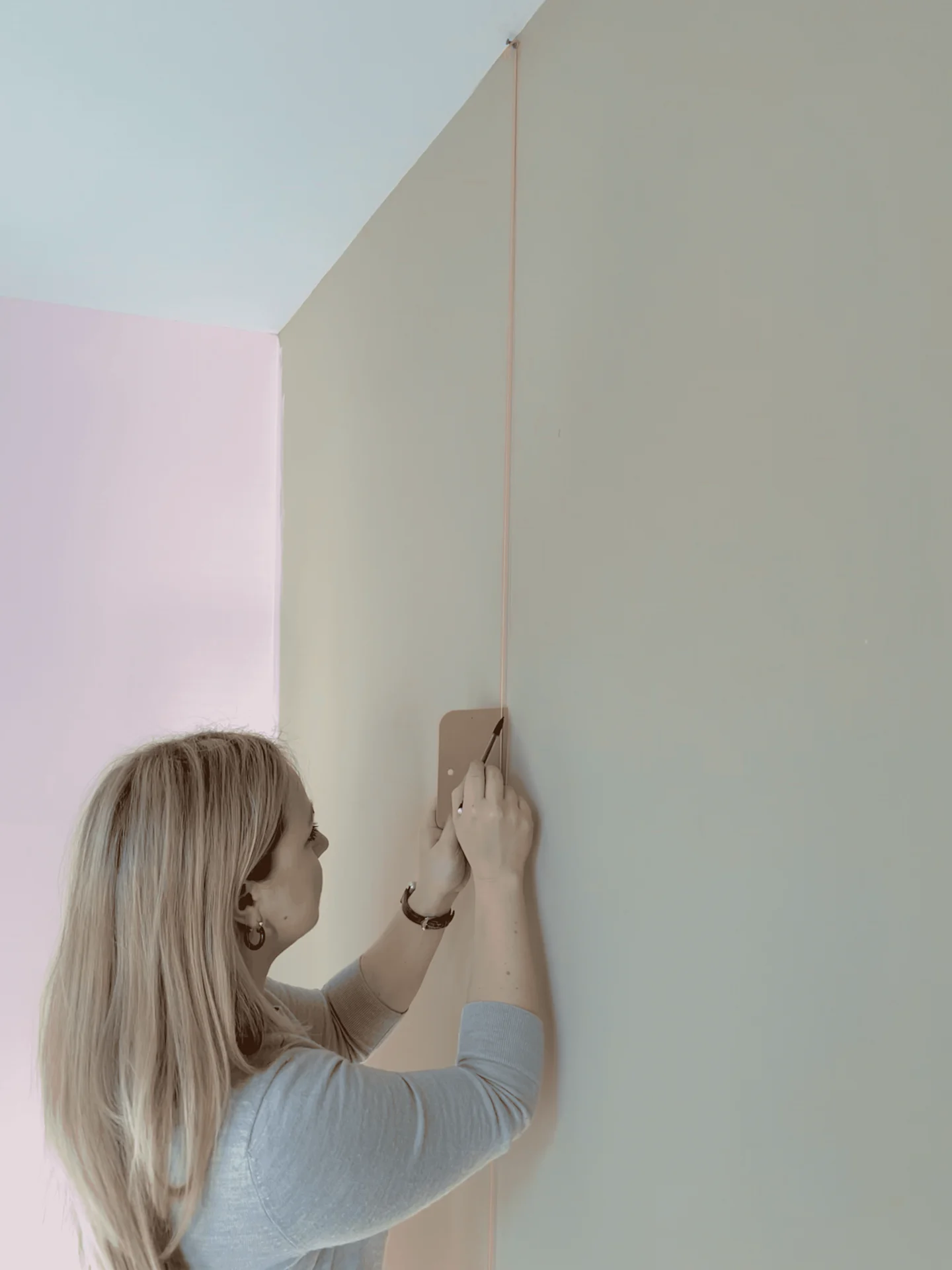 A woman uses a plumb bob to draw a straight line on a beige wall before installation wallpaper.