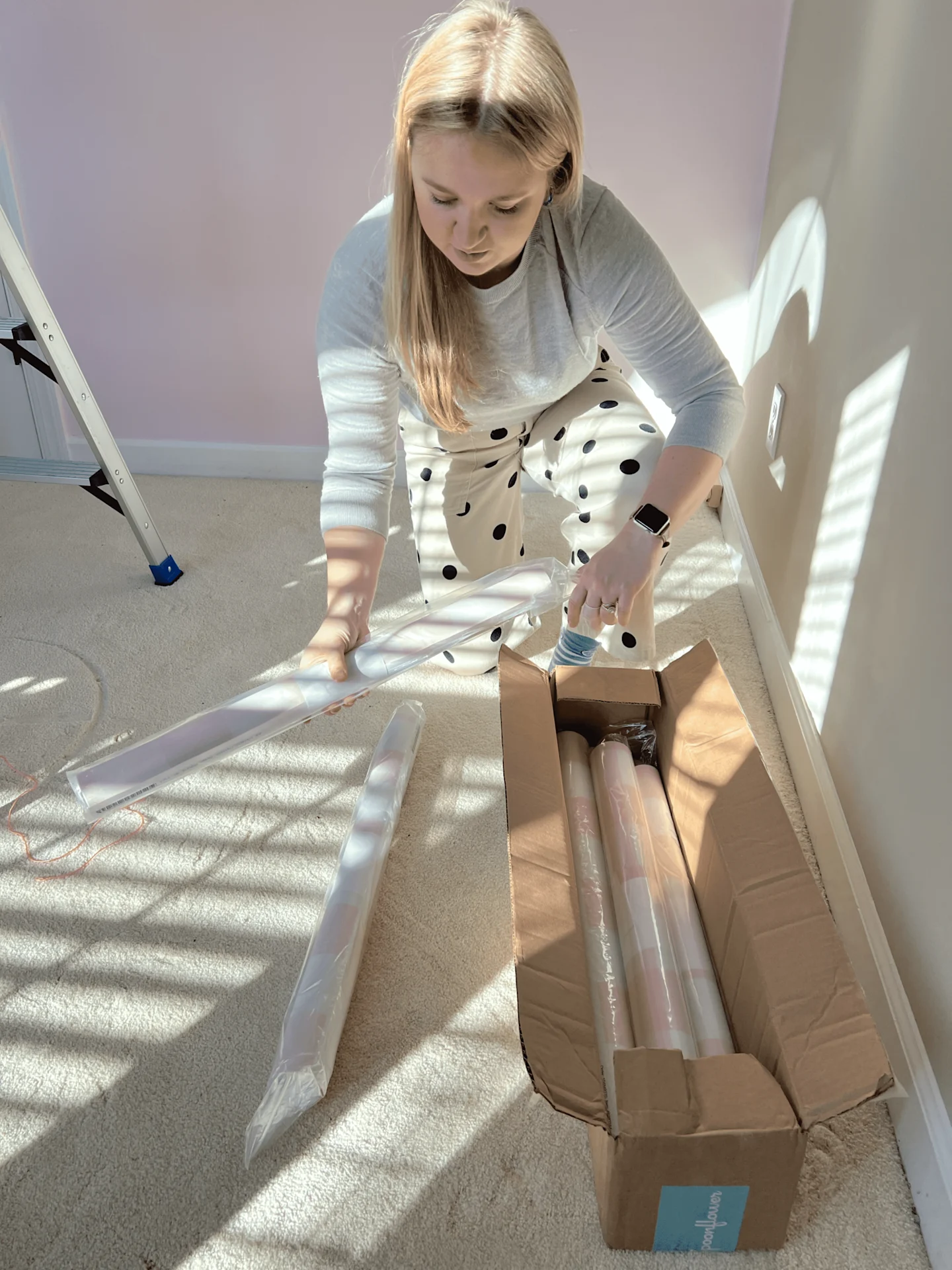 A woman is bent down looking at wallpaper rolls that she is taking out of a box full of them. The wallpaper has a pink-and-white gingham design.