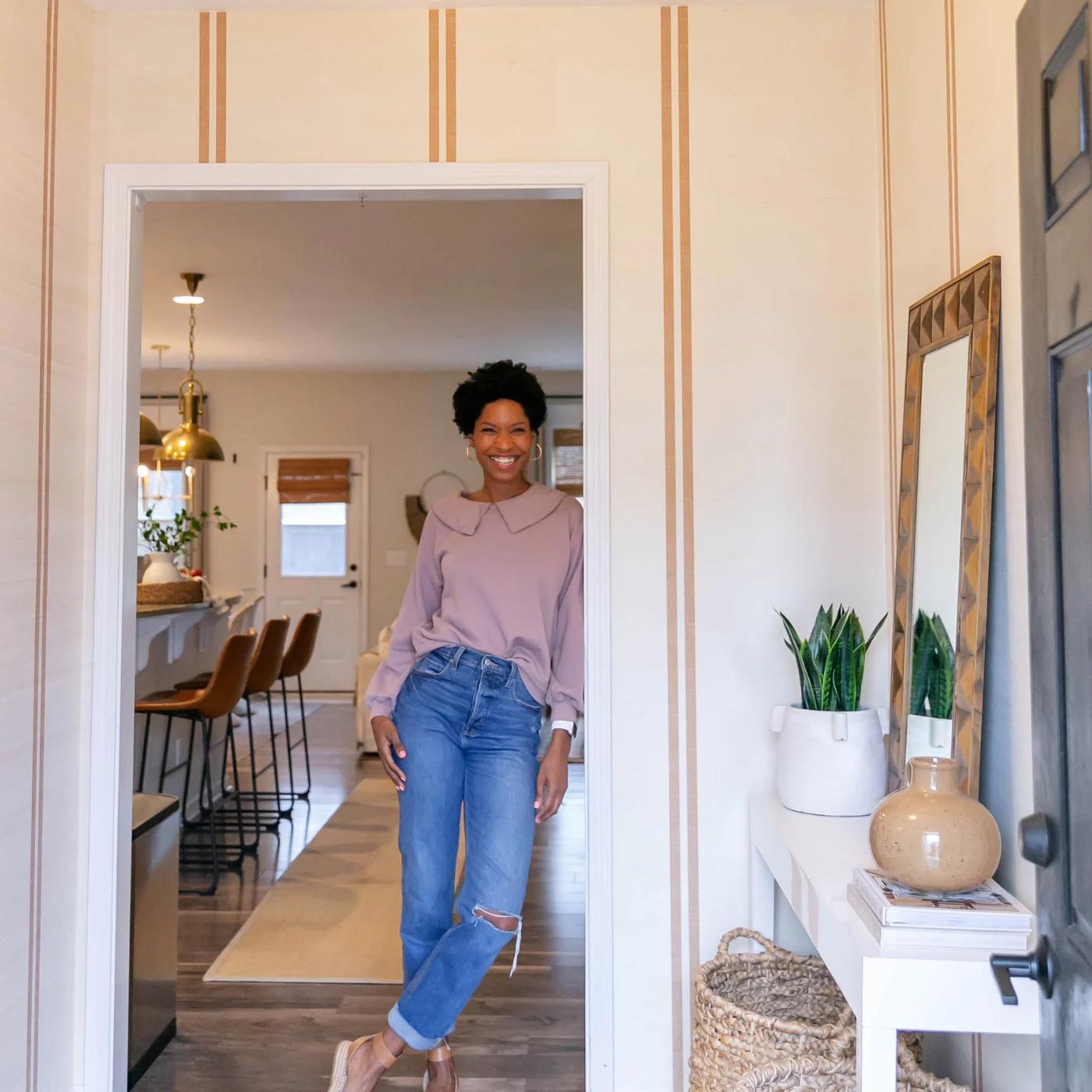 Tiffany stands in the new entryway in her home and looks at the camera smiling. She is wearing a mauve blouse tucked into blue jeans and tan sandles. The design on the wallpaper has a cream background and two small dark tan repeating stripes. To the front