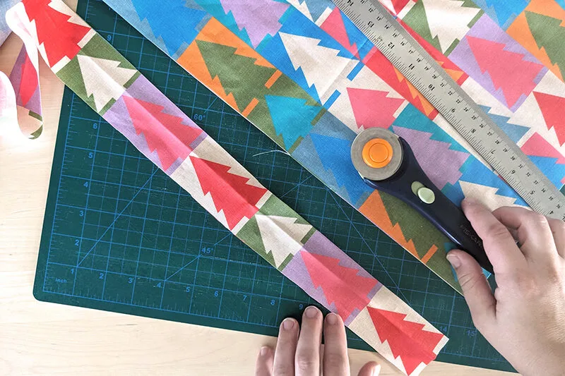 Lisa has cut one strip of fabric lengthwise in a long 2” strip. The fabric features a design with white and jewel tone repeating Christmas trees. The fabric is on a green cutting mat and Lisa is holding a rotary cutter at the photo’s bottom right.