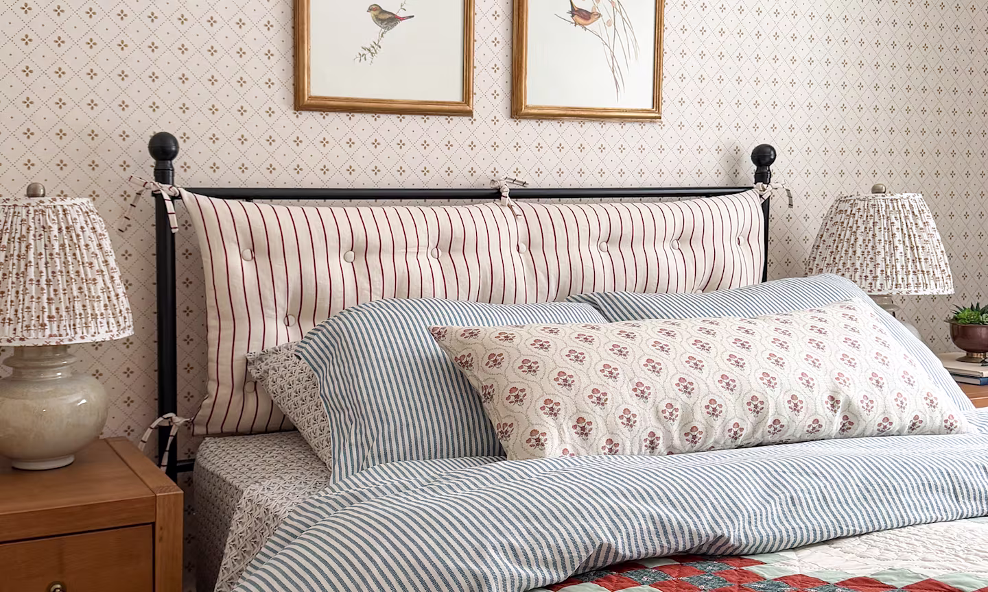 Bedroom with a striped fabric headboard blue pinstripe sheets a block printed fabric lampshade and neutral diamond wallpaper.