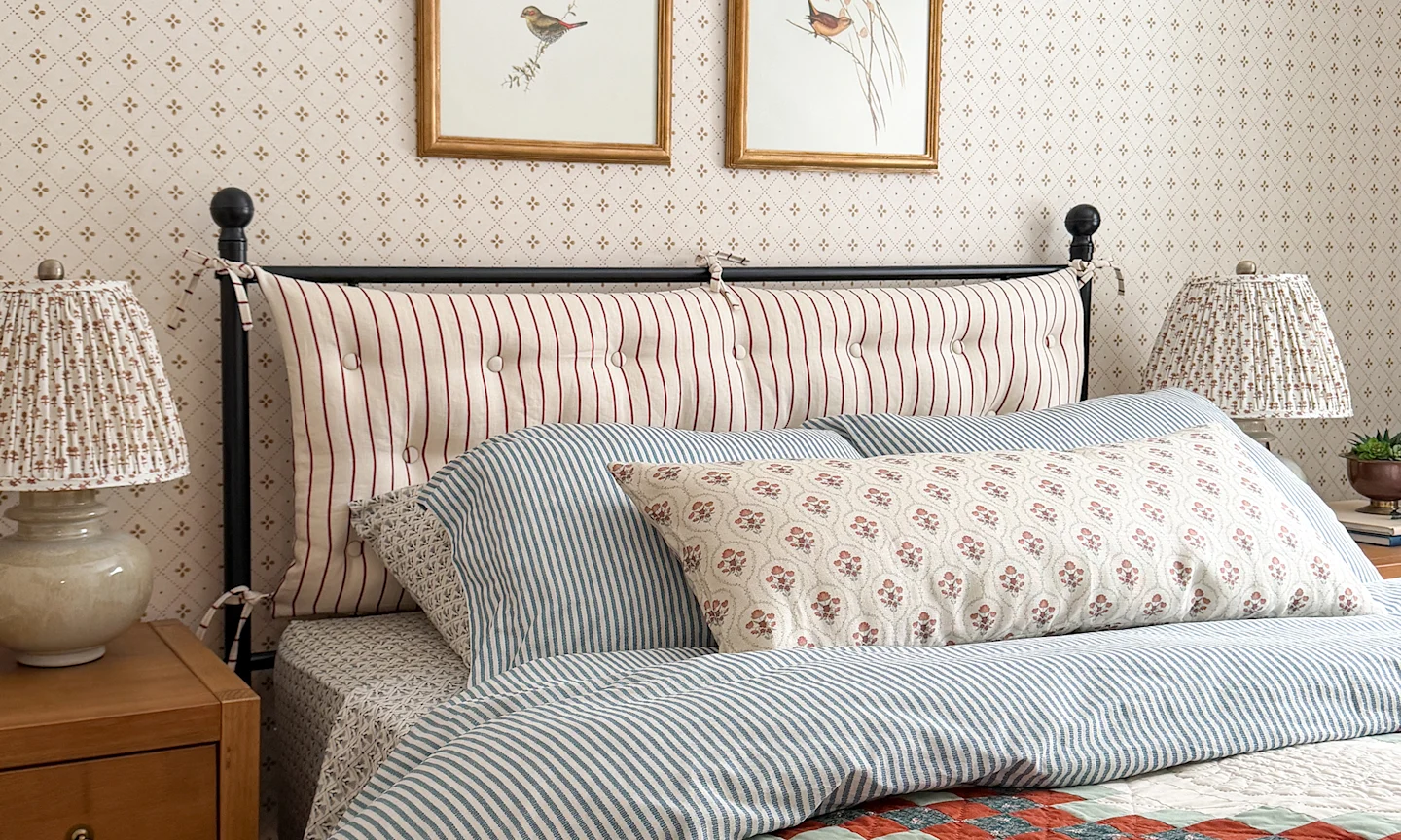 Bedroom with a striped fabric headboard blue pinstripe sheets a block printed fabric lampshade and neutral diamond wallpaper.