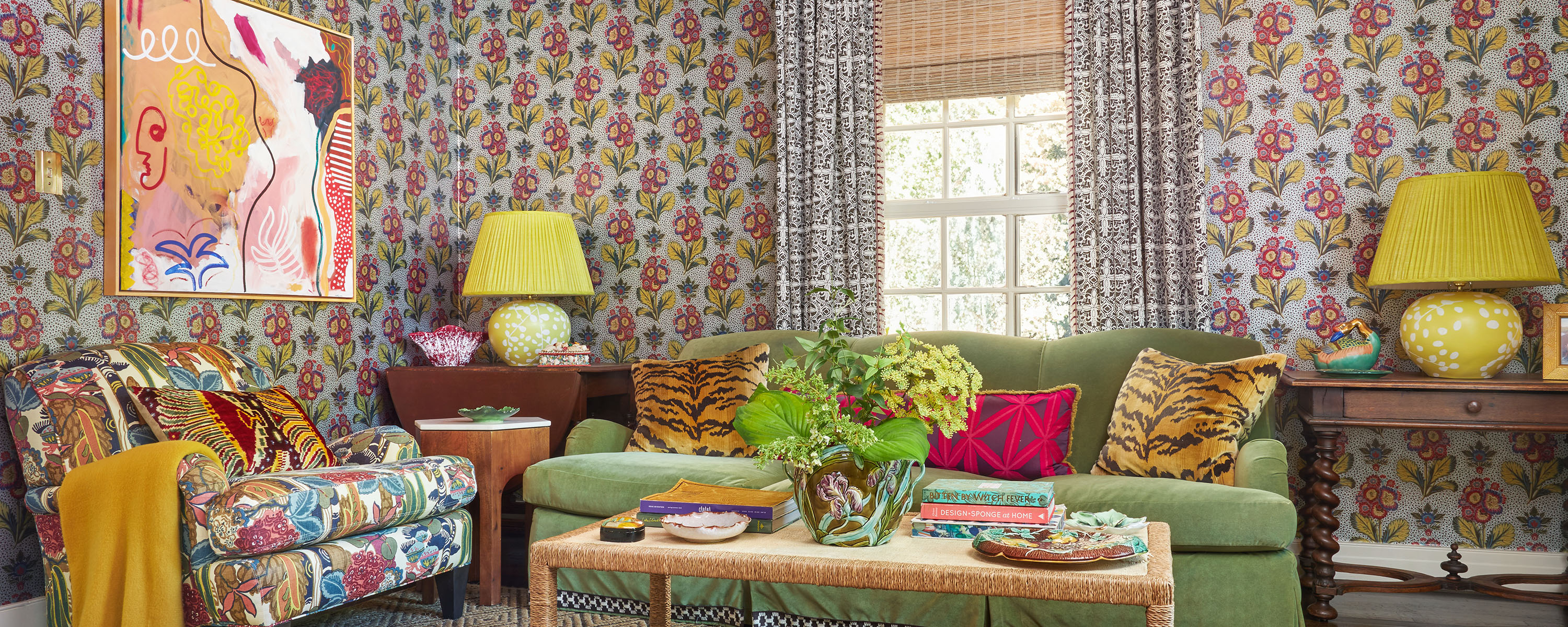 A maximalist living room with metallic floral wallpaper, colorful couches, pillows, lamps, and tables.