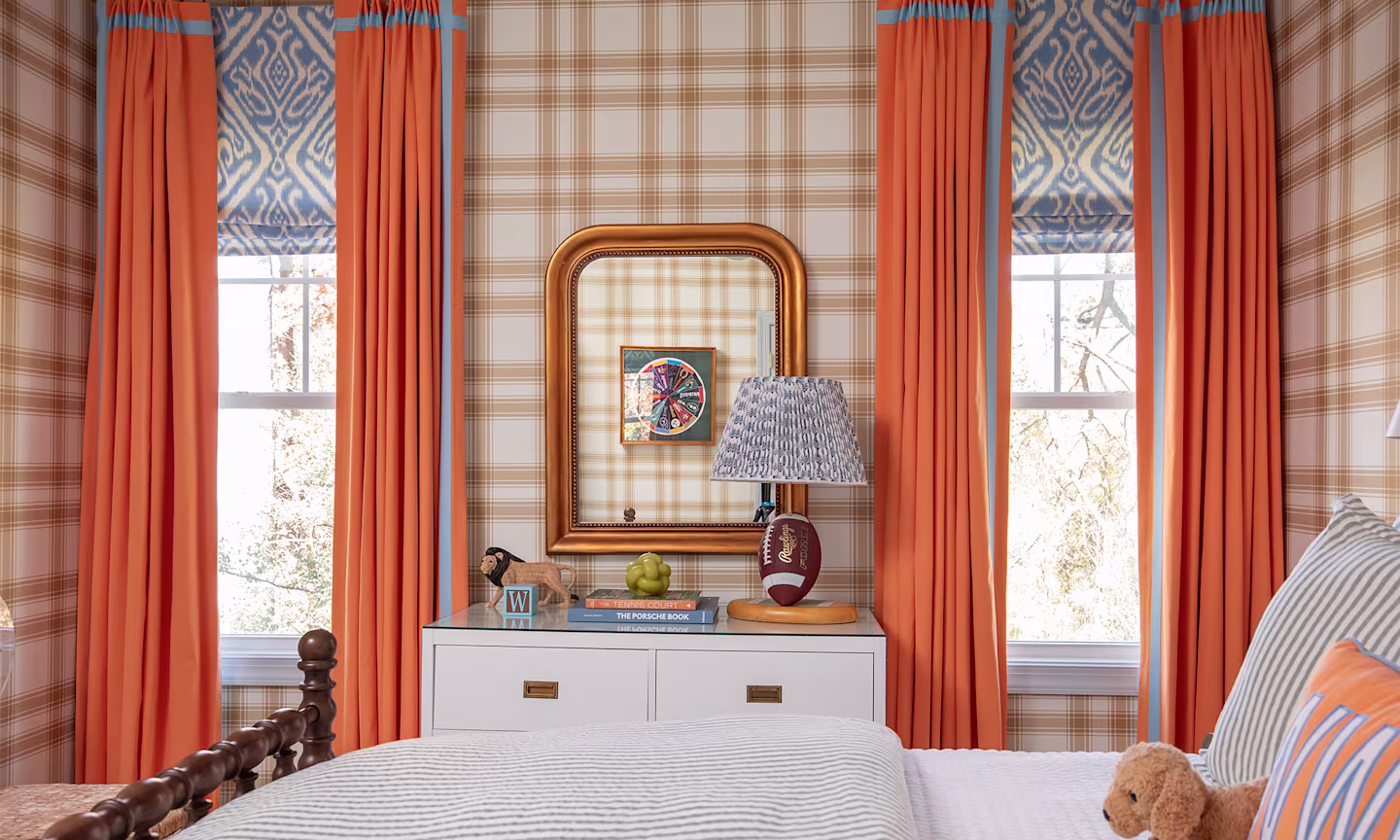 Boys room with a tan and cream large scale wallpaper, blue bedding and orange curtains.