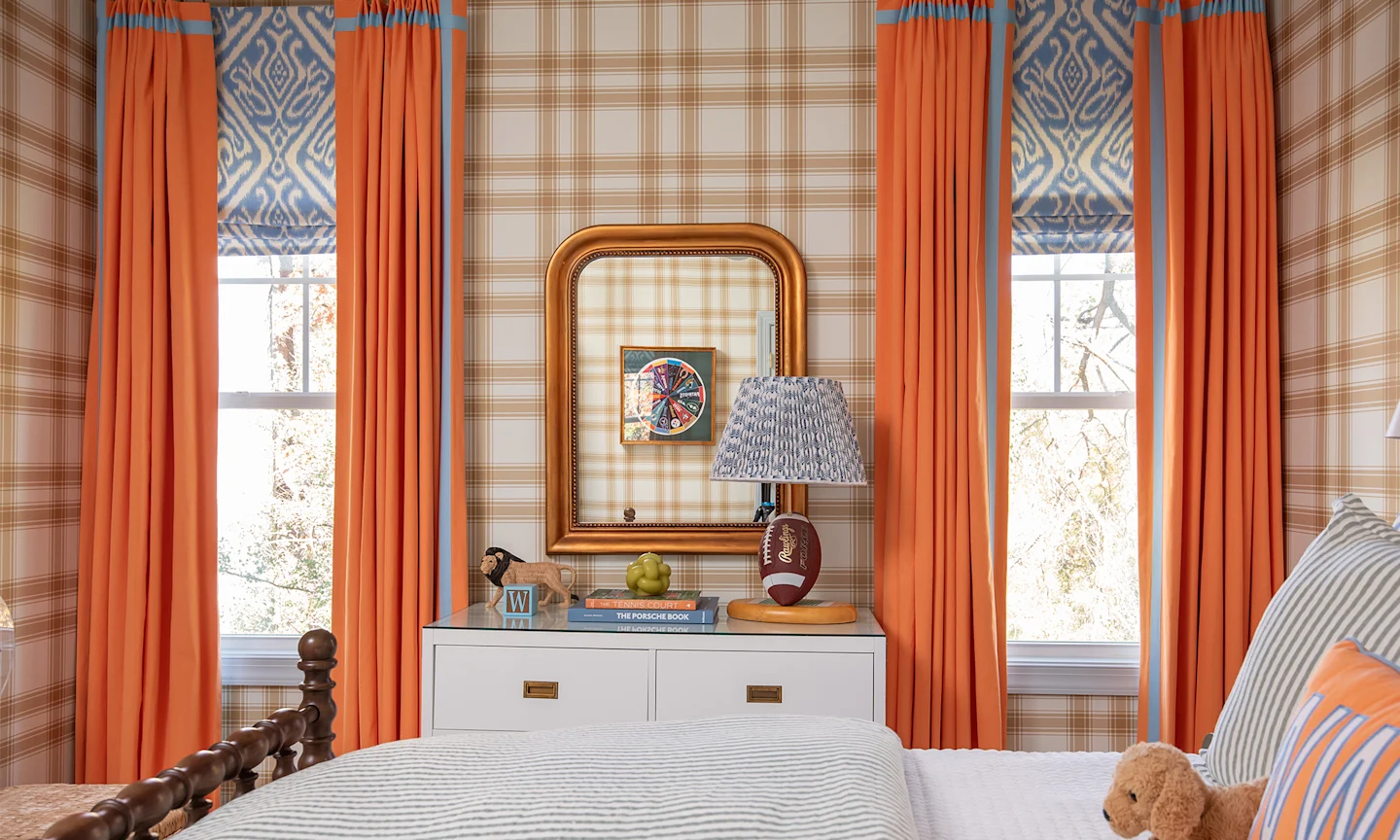 Boys room with a tan and cream large scale wallpaper, blue bedding and orange curtains.