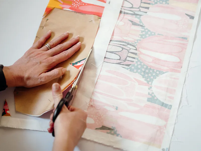 Cutting out the bowl cozy paper pattern that has been pinned and placed on the fabric fold. It is being cut out along the selvage. The fabric design features bright flowers in pinks, reds, yellows and white.