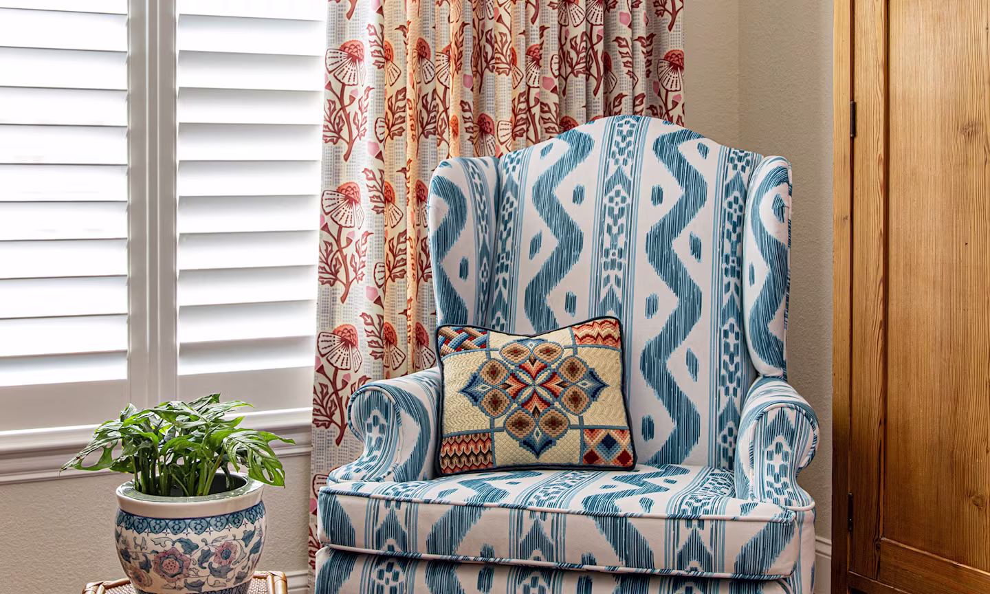 Armchair upholstered in a blue ikat stripe fabric with a pink floral block print curtain on the window behind it.
