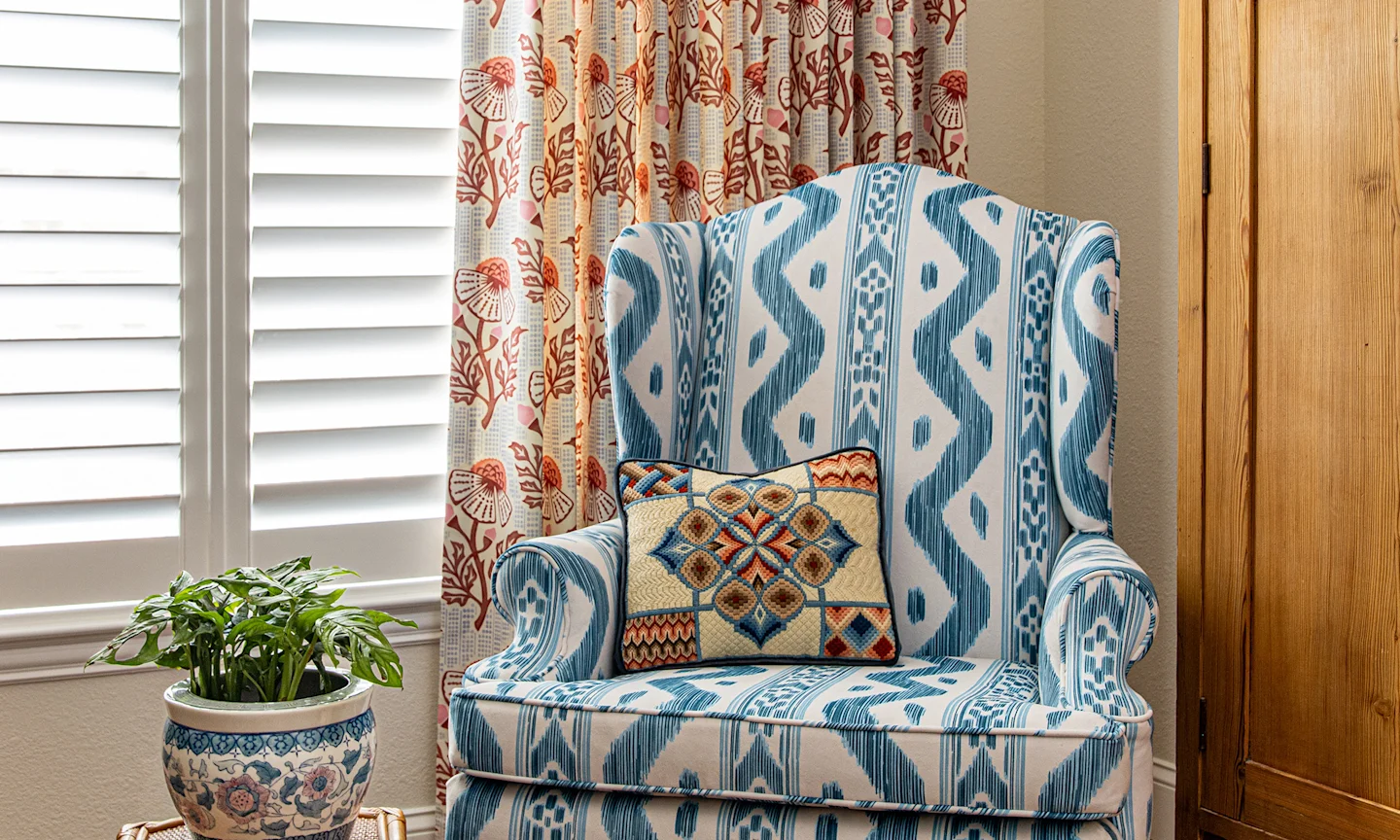 Armchair upholstered in a blue ikat stripe fabric with a pink floral block print curtain on the window behind it.