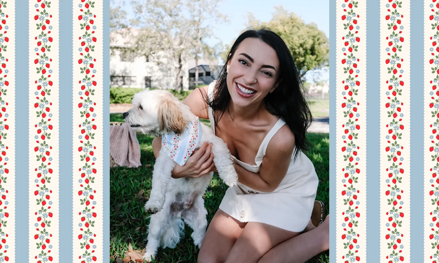 a woman is kneeling on the grown, holding a small dog's front paws up off the ground. the dog is wearing a bandana with stripes and strawberries on it. 