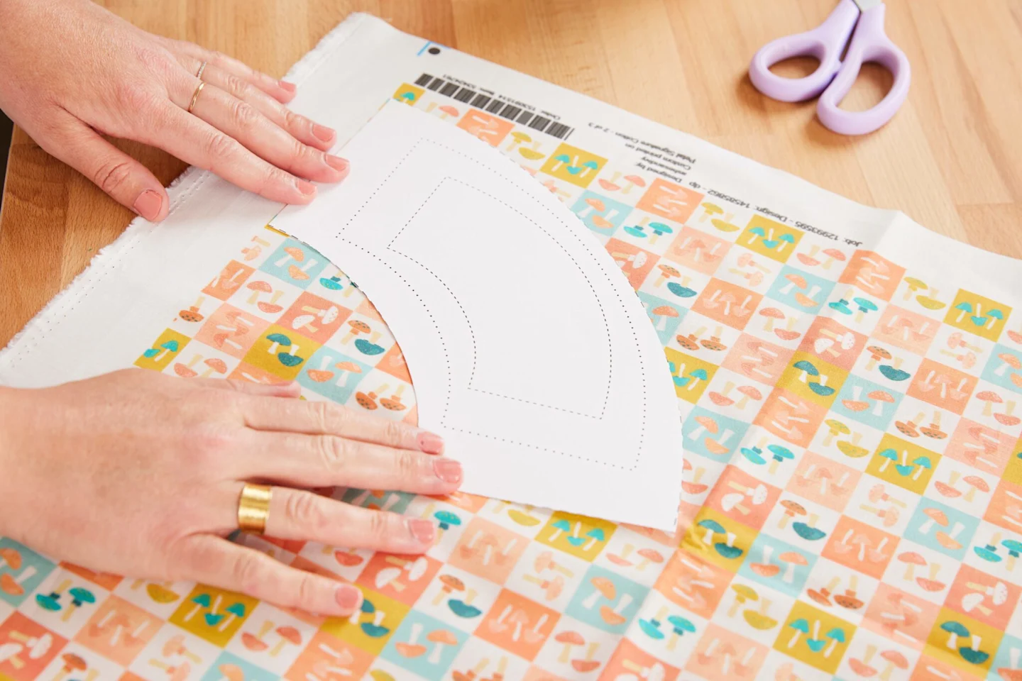 Alexa places the cut-out pattern piece for a 4” (10.2 cm) flower pot from our tutorial template on top of fabric that features small squares each featuring small mushrooms in blue, white, green and peach is laying on the table.