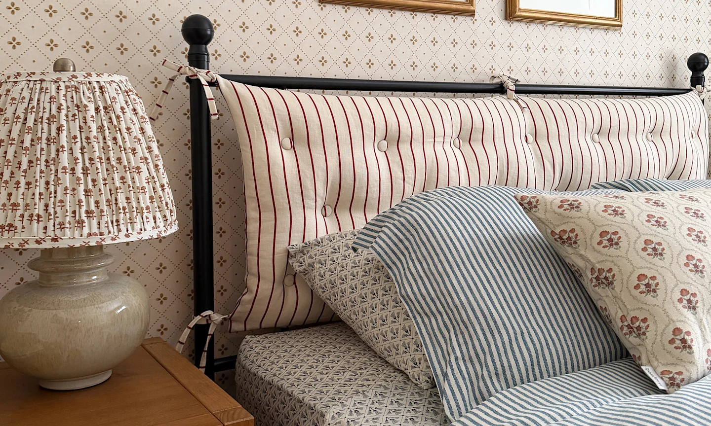 Bedroom with a striped fabric headboard blue pinstripe sheets a block printed fabric lampshade and neutral diamond wallpaper.