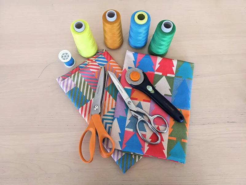 Materials for this project have been laid out on a wooden table: thread spools in yellow, orange, blue and green; two folded pieces of cotton fabric featuring designs in jewel tone colors, in both a Christmas tree repeating design and small squares of coo