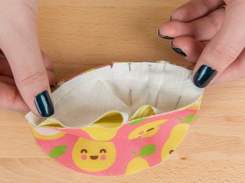Two hands are holding the baby shoe toe pieces that are sewn so far. Sewing pins indicate the other half that needs to be sewn.