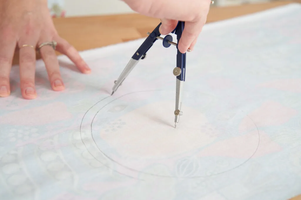 An image of a compass being turned to draw a second circle on the wrong side of a piece of fabric. This second circle is larger and slightly outside the first circle. 
