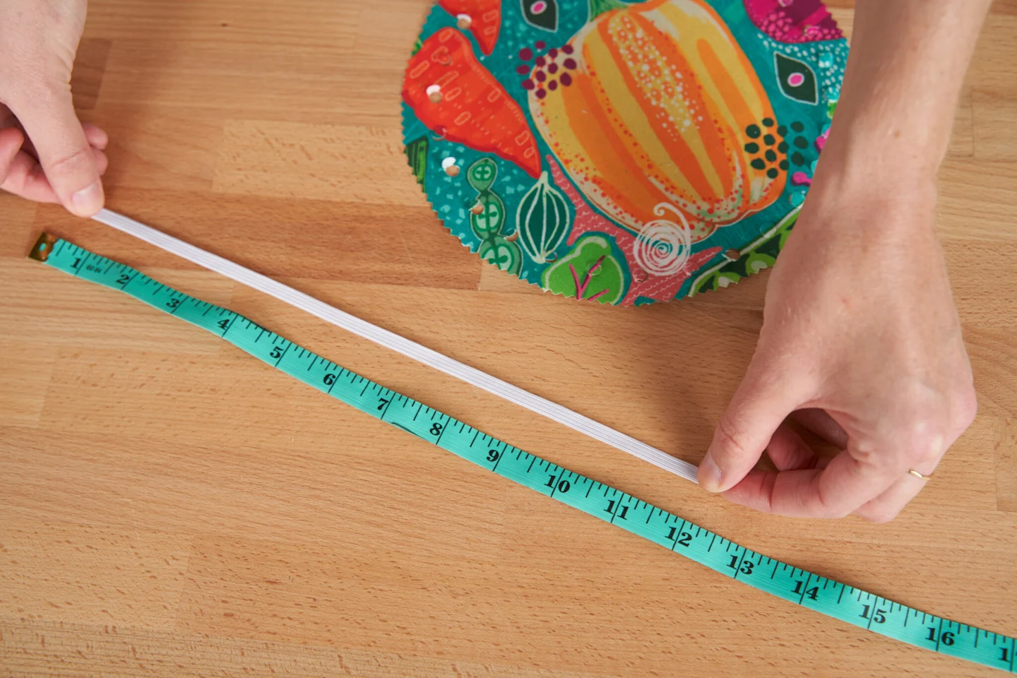 A person uses measuring tape to measure a width of elastic. A piece of fabric which has been cut into a circle and had holes punch near the edge, lays toward the top of the photo. The design on the fabric has a teal background and vegetables, a pumpkin, p