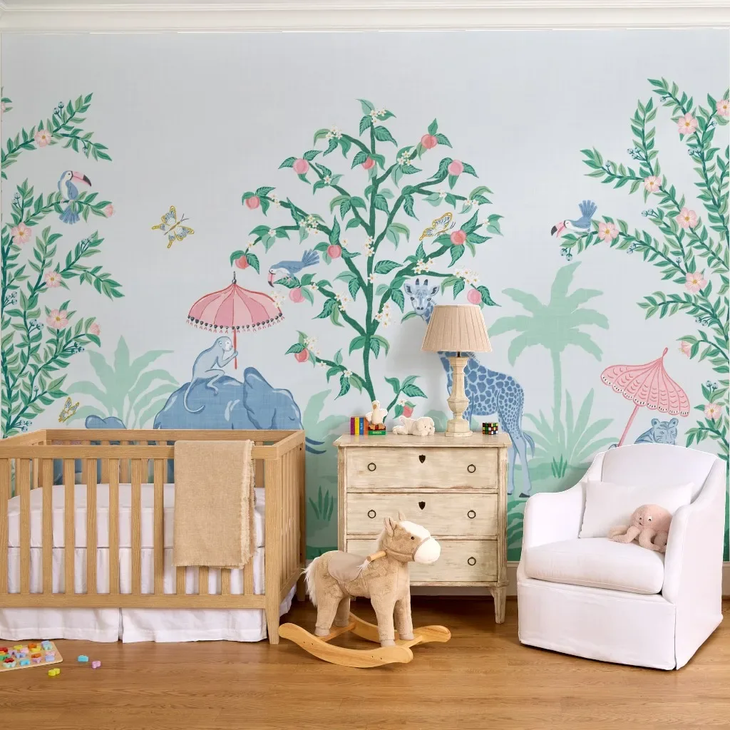 Nursery with a whimsical jungle wall mural featuring giraffes, monkeys, tropical birds, flowering trees, a wooden crib, vintage dresser, rocking horse, and white reading chair.