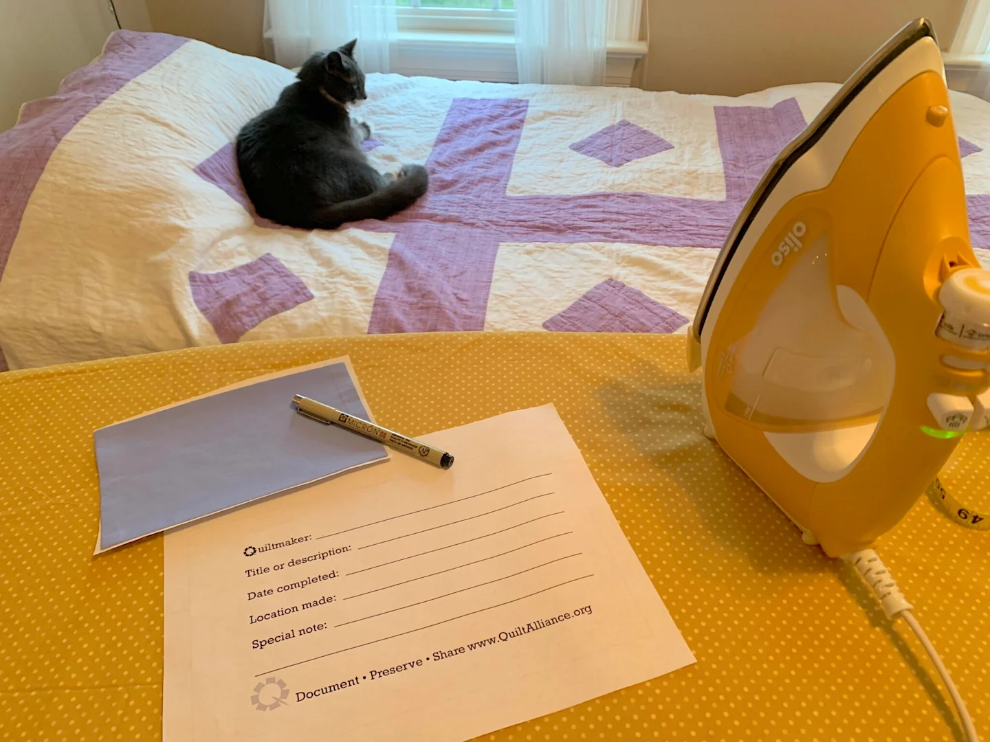 A piece of paper lays on a yellow ironing board next to a black pen. It has the following prompts, each followed by a blank link on which to write the answer: Quiltmaker, Title or description, Date completed, Location made, Special note. Underneath those 