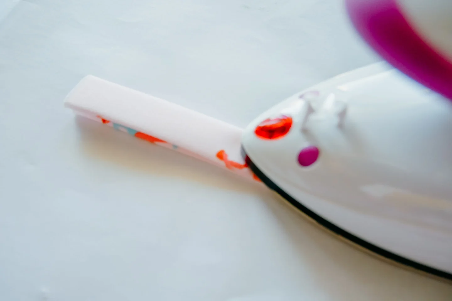 A small rectangle of fabric has been folded together and is being pressed lengthwise with a hot iron. The fabric features a design with small female surfers surfing through a pink background.