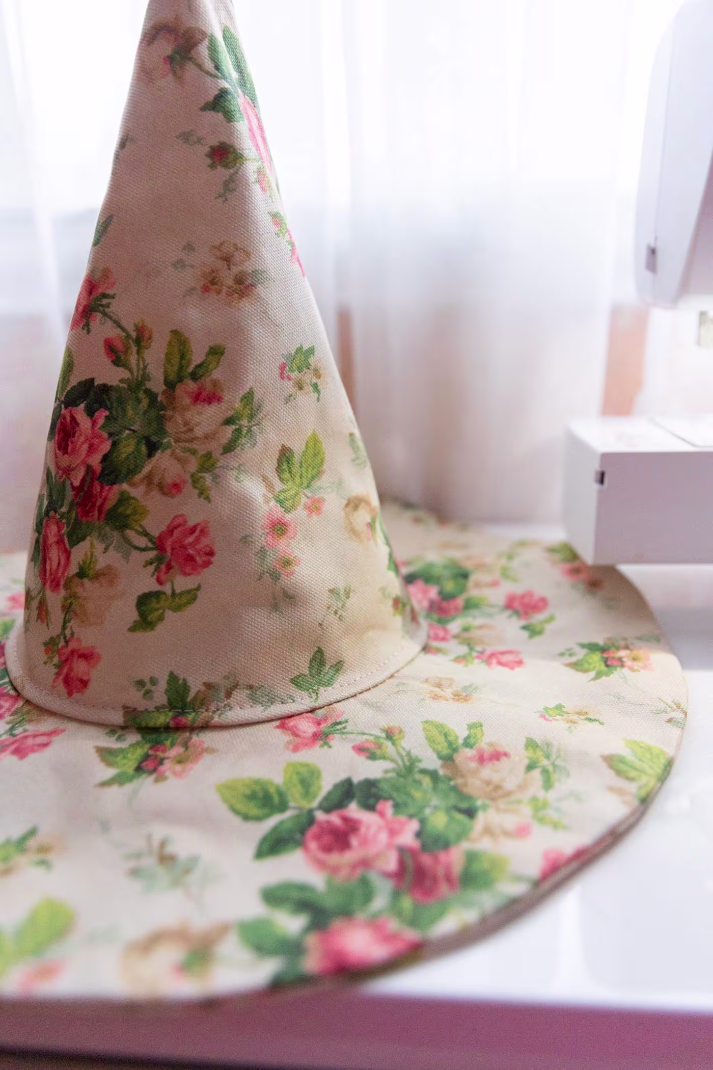 The finished hat sits on a white desk beside the sewing machine. A sheer curtain in front of a window is in the background.
