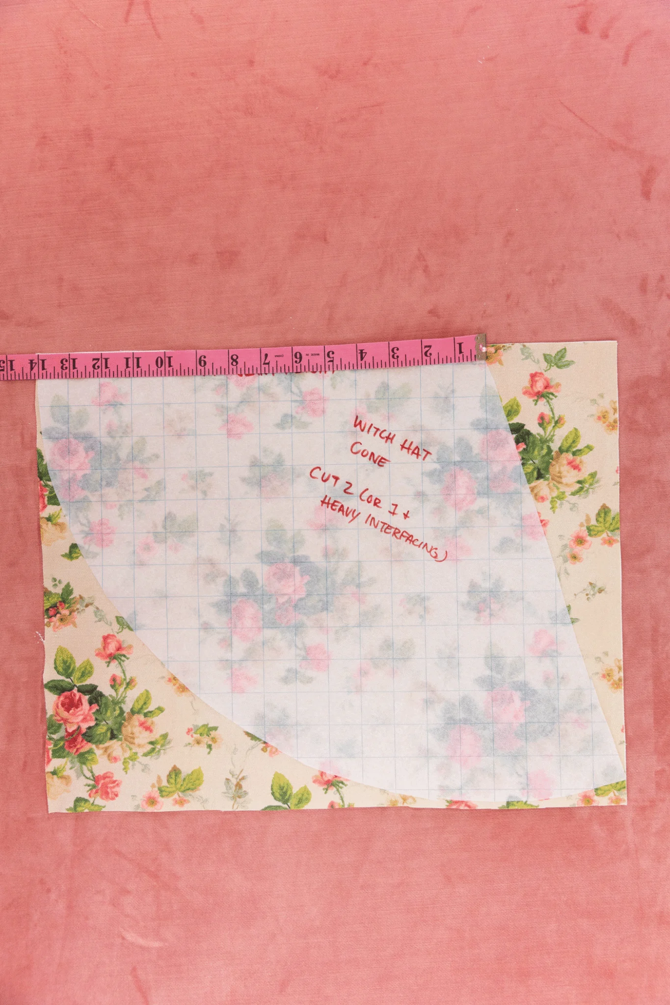 A translucent grid paper is on top of a cream floral fabric with pink roses and green leaves. One pink measuring tap lays on the edge of the fabric measuring out 14-inches. The background is a pink carpet. The measuring tape moves across the grid paper un