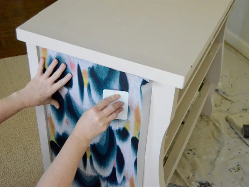 Barb smoothes out the wallpaper that is attached to the side of the dresser with a plastic squeegee.