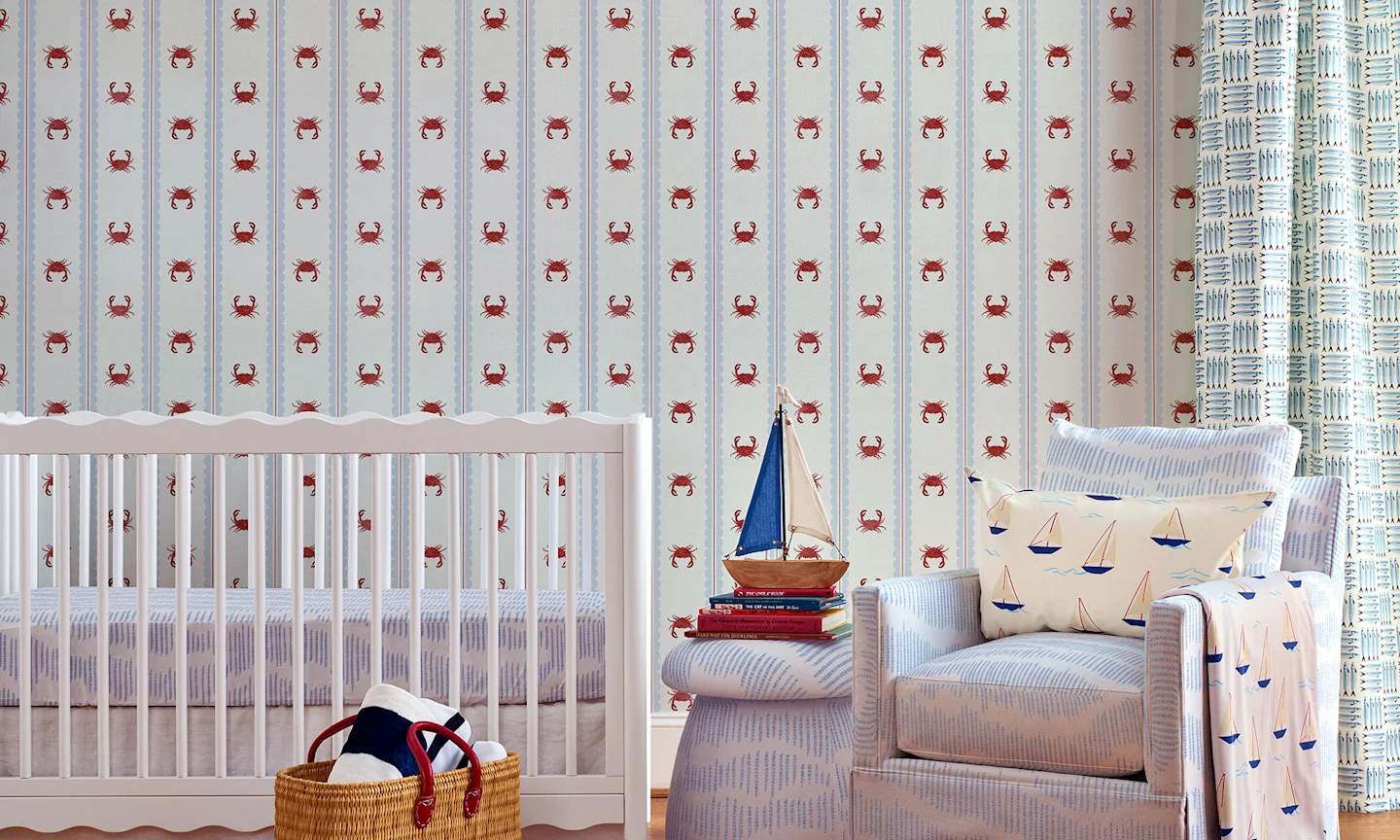 Nursery with striped crab wallpaper, blue upholstered chair, ottoman and a lumbar pillow and blanket in a sailboat pattern.