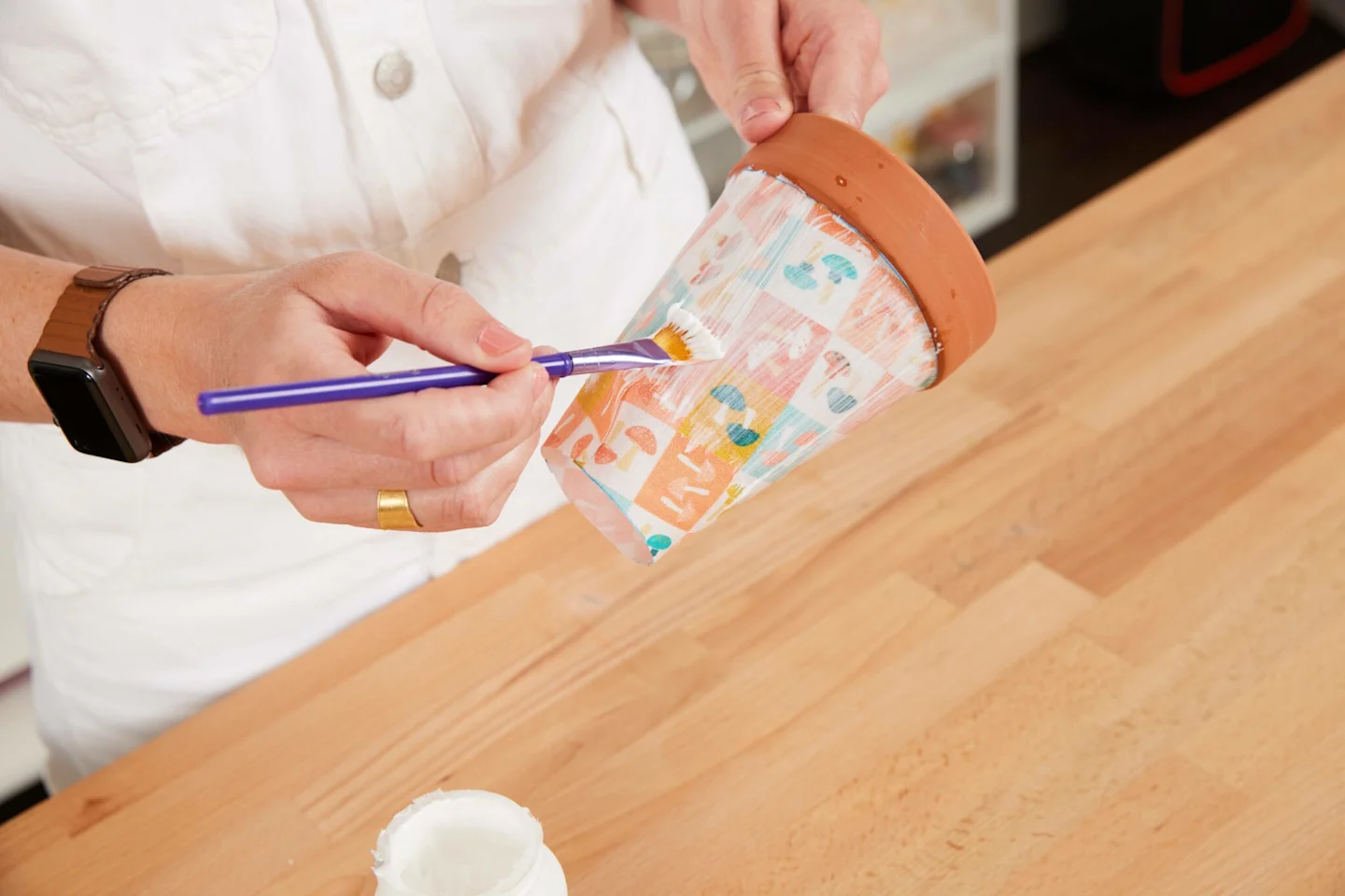 Using a paintbrush, Alexa paints the flower pot covered with fabric featuring small squares of small mushrooms in blue, white, green and peach with Mod Podge Outdoor.