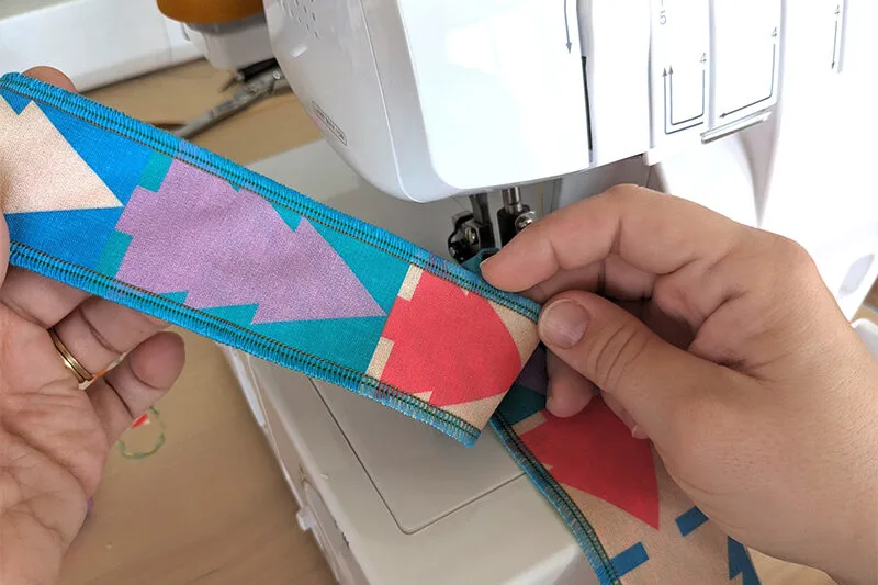 A hand holds up 2 2”-wide strips of fabric that have been serged together along both their right-hand and left-hand side. The strips of fabric have a Christmas tree repeating design, also in jewel tones. A white sewing machine is in the middle of their ha