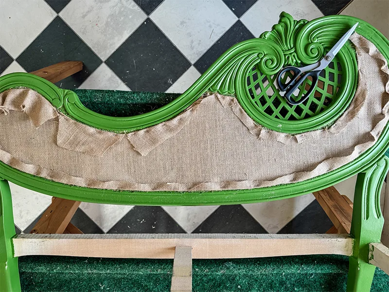 A layer of burlap is stapled onto the jute webbing of the sette top. Scissors sit on a decorative part of the chair's top. A checkered floor is in the background.