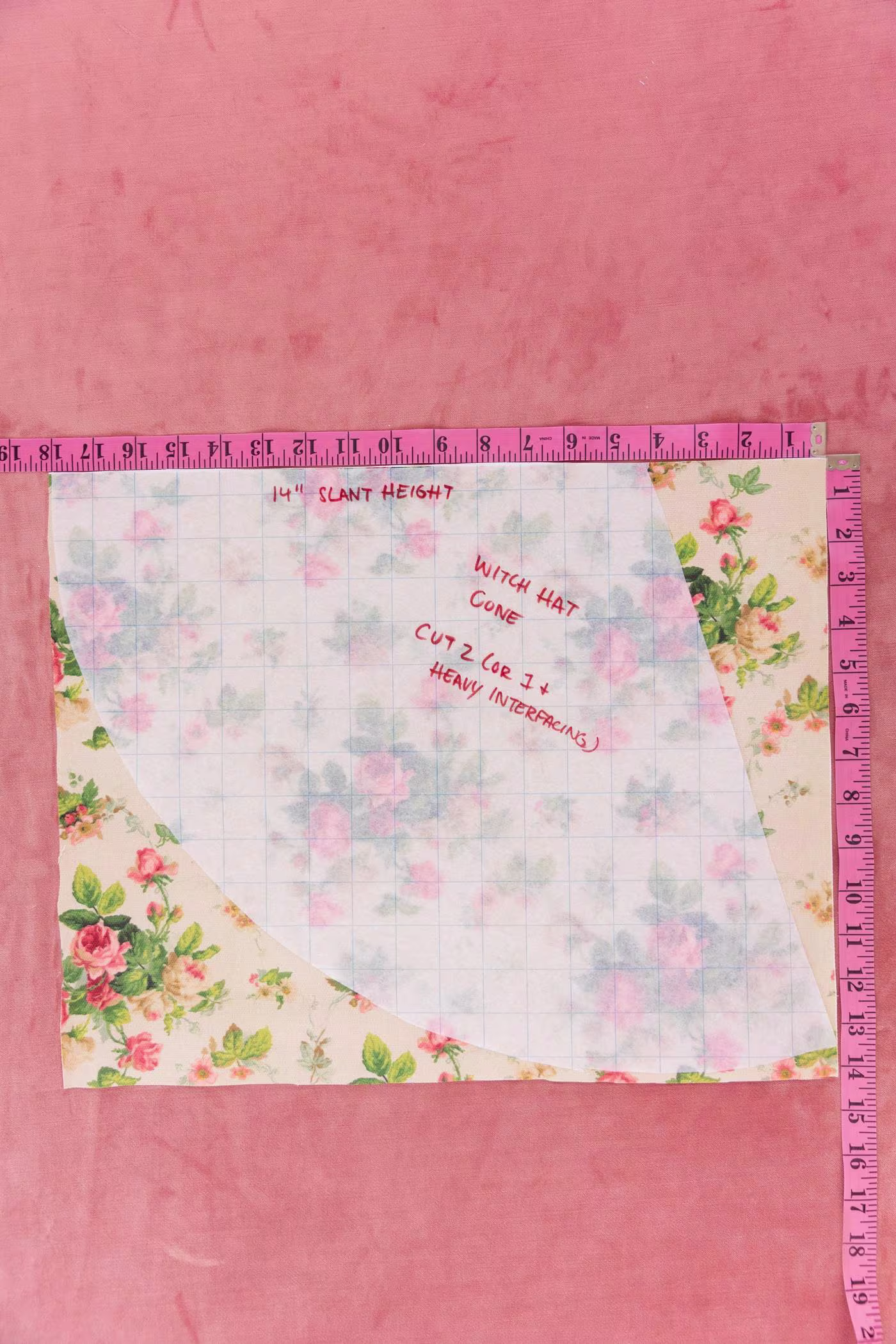 Translucent grid paper lays on top of a cream floral piece of fabric with pink roses and green leaves and stems. Two pink measuring tapes are beside the fabric measuring its length and width. A pink carpet is behind as the background.
