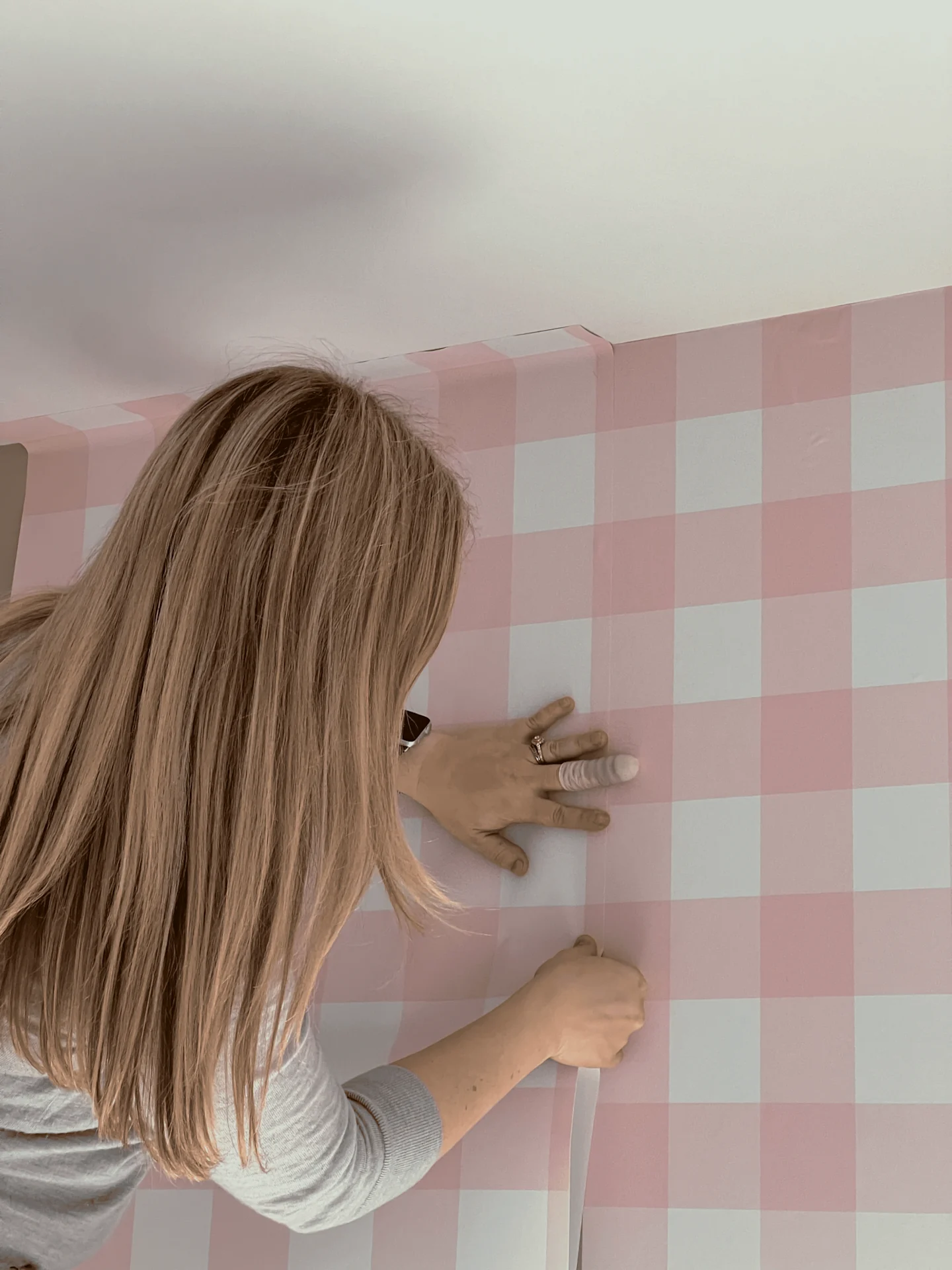 A woman inspects that two rolls of installed wallpaper are lining up down as she installs wallpaper with a pink-and-white gingham design on a wall.