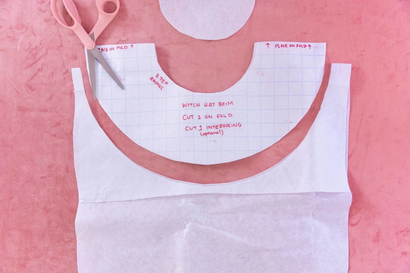 A peach-colored pair of scissors rests on the semi-circle template used to make the brim. Excess heavy interface paper is a few spaces apart from the template.
