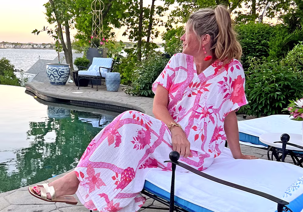 Danika sits by a pool in a V-neck caftan with hot pink flowers on a white background.