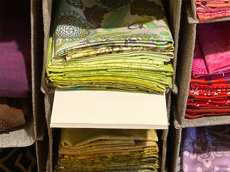 Stacks of green printed fabric are folded in a clothing organizer with a piece of foam beneath the last fabric piece. Other colored fabrics are in other surrounding organizers.
