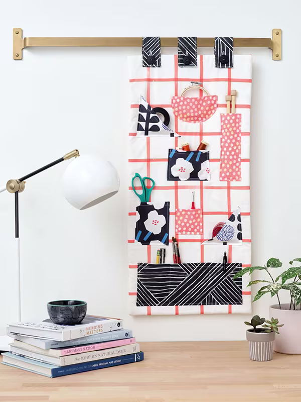 A fabric craft organizer hangs from a wooden dowel attached a wall. It has small pockets, each filled with a different craft tool, including tape, scissors, spools of thread and more. It hangs above a desk with small plants, books and a white lamp.