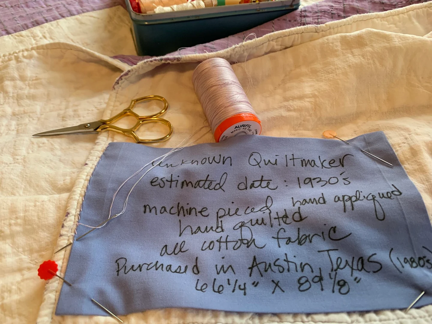 A blue piece of fabric has been stitched to the white back of a quilt. The fabric is pinned to the back on the quilt and the stitching in place is in progress. A small pair of scissors and a spool of light gray thread lay at the top of the label. On the q