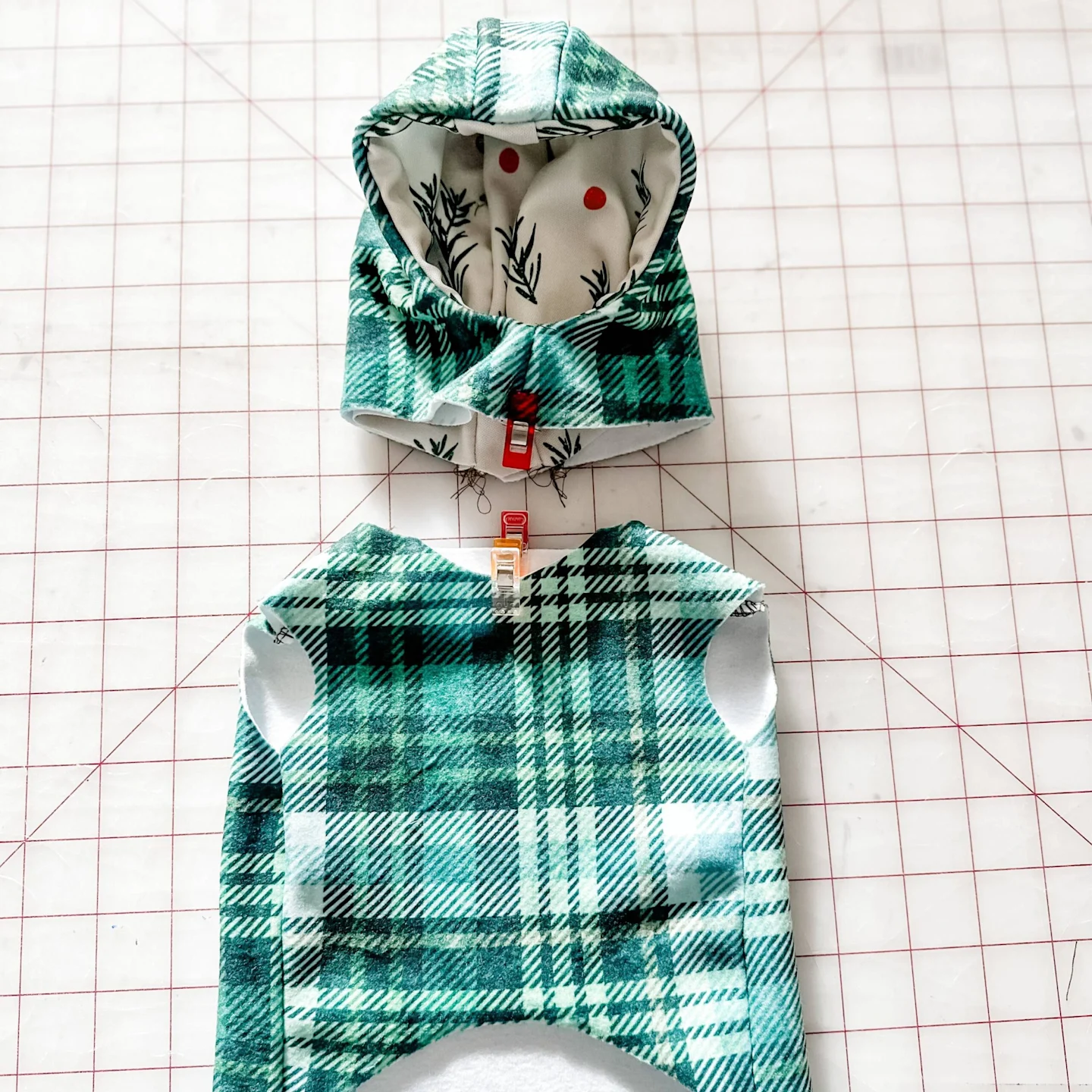 A Plaid green hoodie with green, white and red interior has sewing clip securing the bottom of the hood piece. A sewing clip is clipped to the center of the Green plaid bodice below the hoodie. The clips are lined up vertically. A grid surface is in the b