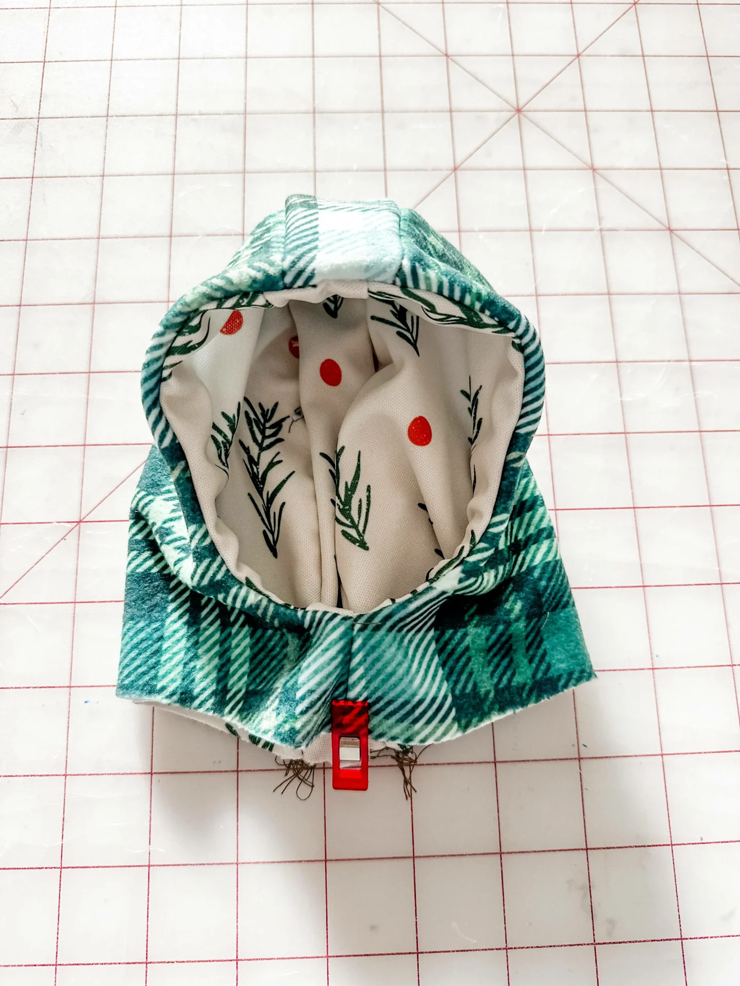The hoodie is flipped so the exterior, green plaid design is facing up. From the inside the interior, white green and red design is also visible. A sewing clip secures the bottom center of the hoodie where the bodice will soon be connected. A grid surface