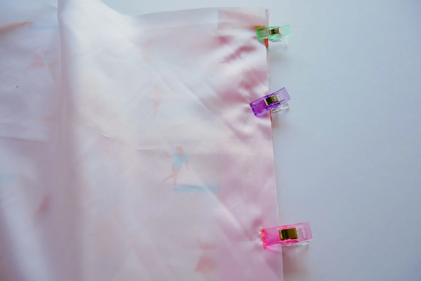 A rectangle piece of Polyurethane Laminate fabric is lying on top of a rectangle of fabric featuring a design with small female surfers surfing through a pink background. A pink zipper lays in between the two. Pastel wonder clips have been placed along th
