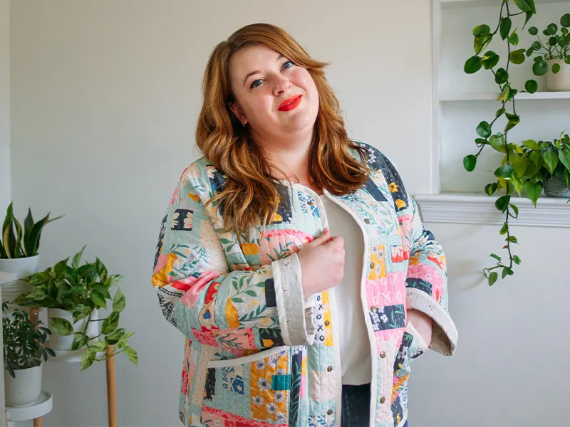 A close up of Meg's cheater quilt quilted jacket featuring delicate florals, most with a blue or pink theme. She is standing in front of a white wall.