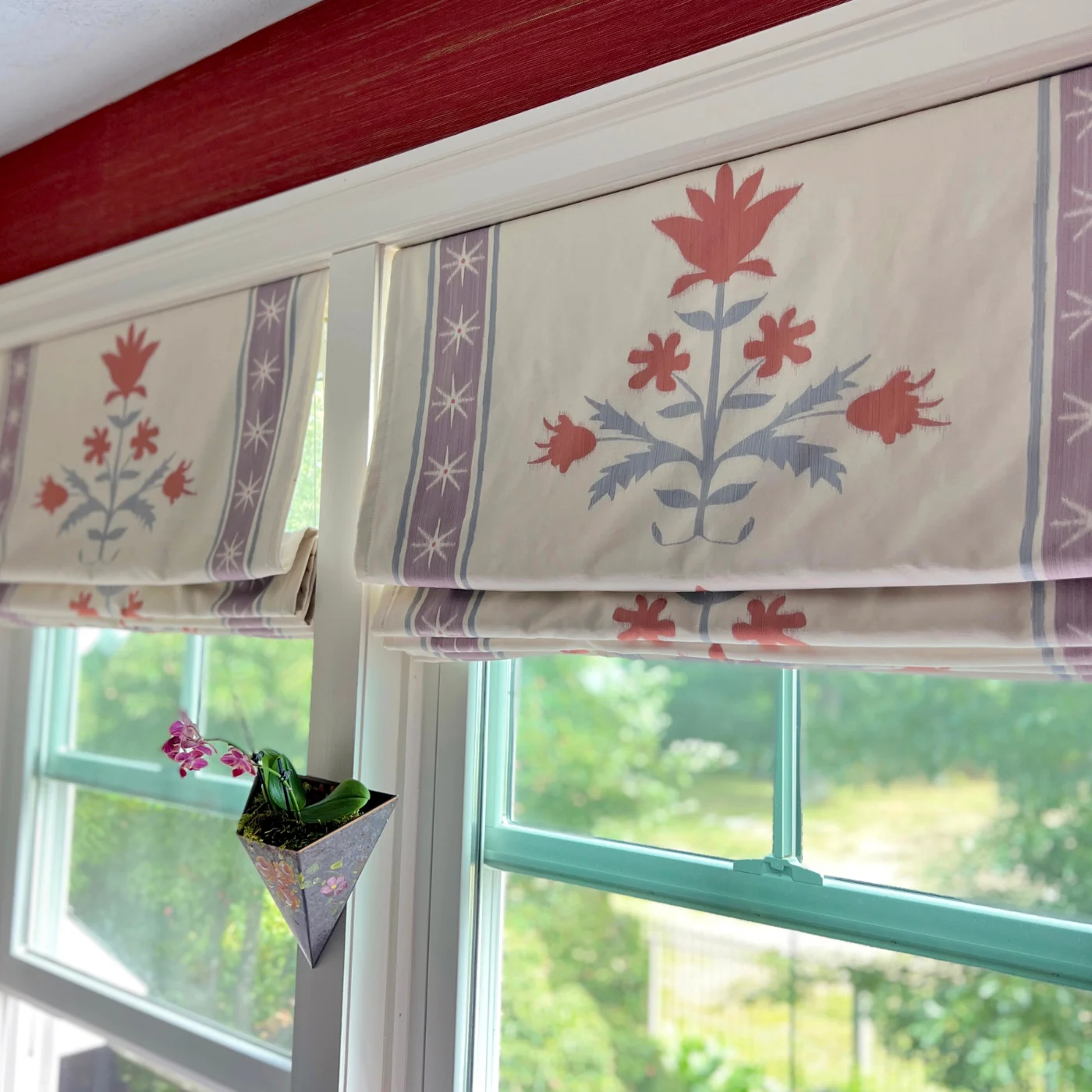 Two Roman shades sit side by side on two windows. The shades have what looks like a band at the edge featuring a thick purple stripe with white stars. The stripe has thin blue stripes on either side of it. The band is actually part of the design! The midd