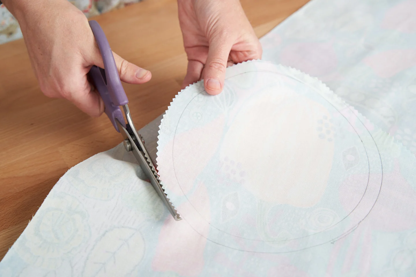A person is using pinking shears to cut around the outside edge of the second, larger circle drawn onto the wrong side of fabric. 