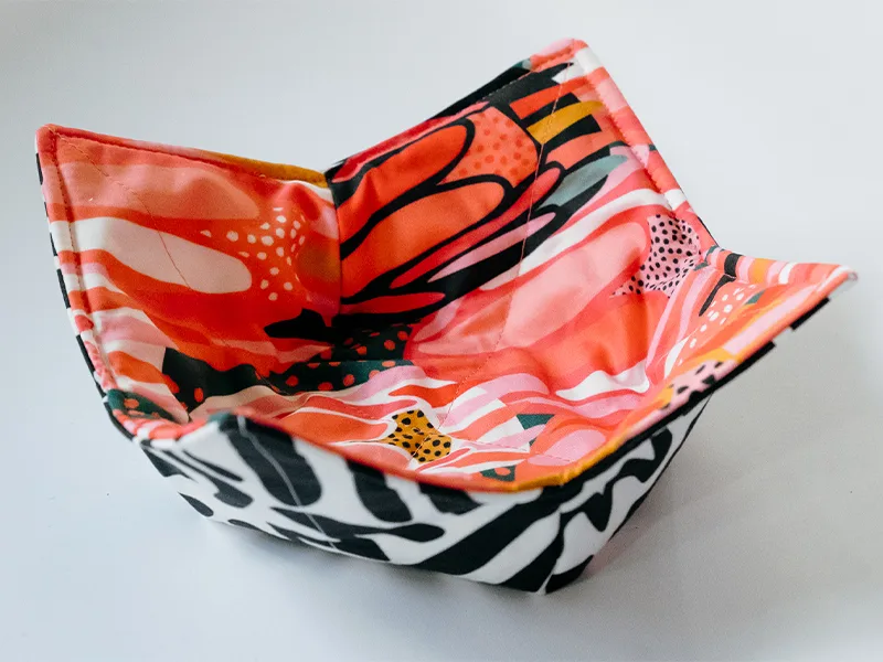 The finished bowl cozy as seen from the right side. It has a concave shape to fit a bowl of soup and four pointed edges. The fabric on the top side features bright flowers in inks, reds, yellows and white. The fabric on the bottom side features black geom